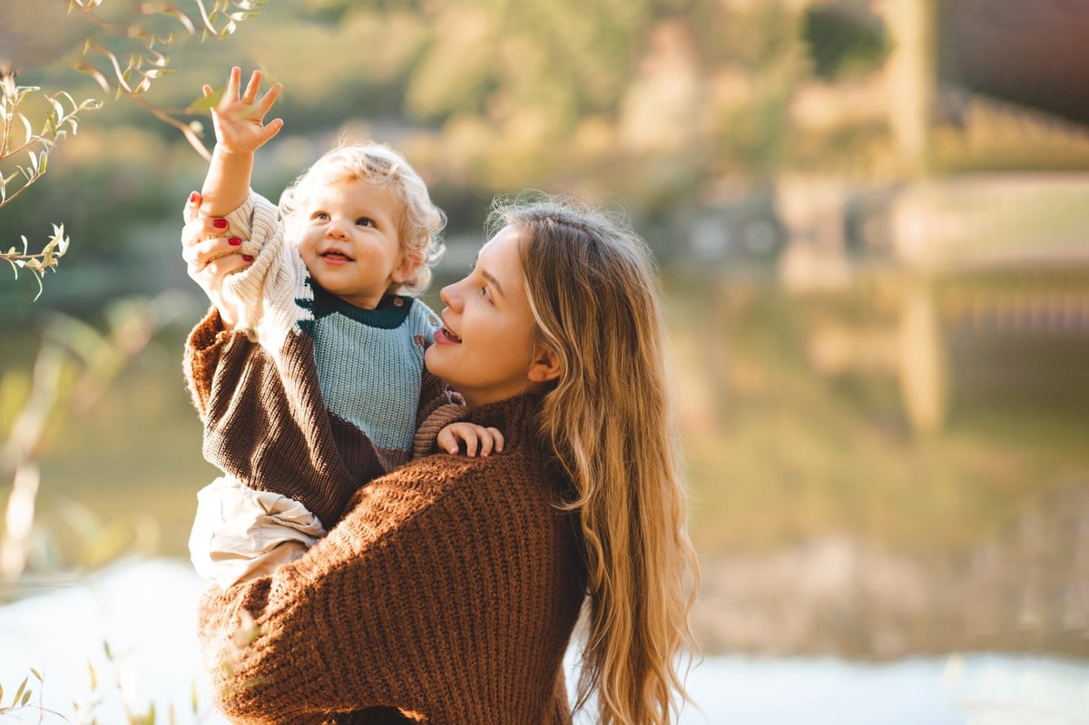 Smiling mother holding baby girl over lake and nature background, warm autumn scene